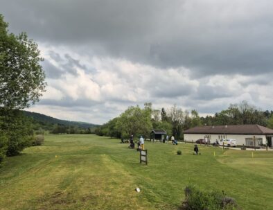 Vue panoramique d'un terrain de golf verdoyant sous un ciel nuageux, avec des golfeurs utilisant des chariots sur le parcours. En arrière-plan, une maison de club et des arbres luxuriants ajoutent à l'atmosphère paisible. L'image illustre un cadre idéal pour les amateurs de golf, combinant nature et sport dans un environnement relaxant.