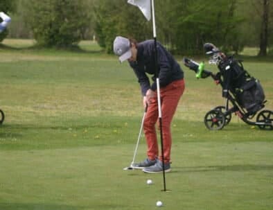 Un golfeur s'apprête à putter sur le green, concentré sur sa frappe. En arrière-plan, une golfeuse observe, tandis qu'un chariot de golf est stationné à proximité. L'image illustre une belle journée de golf en plein air, mettant en avant l'équipement et l'ambiance conviviale du sport.