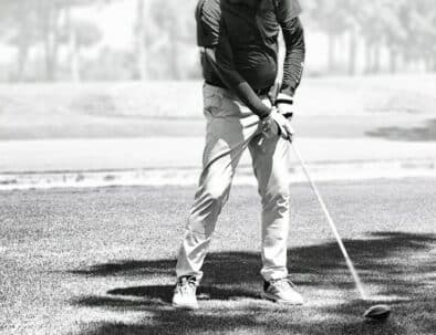 Un golfeur se prépare à frapper une balle sur un parcours de golf verdoyant. Il porte une tenue de golf adaptée, comprenant un polo et un pantalon clair, ainsi qu'une casquette pour se protéger du soleil. L'image en noir et blanc met en avant la concentration du joueur, tandis que l'arrière-plan présente des arbres flous, créant une atmosphère calme et professionnelle. Cette scène illustre la passion et la précision requises dans le sport du golf.