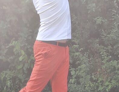 Homme en train de jouer au golf, effectuant un swing dynamique avec un club. Il porte un t-shirt blanc et un pantalon orange, se tenant sur une pelouse verdoyante. En arrière-plan, on aperçoit des buissons verdoyants. Un ballon de golf aux couleurs vives est visible au sol, près de ses pieds. Cette image illustre l'activité de golf en plein air, mettant en avant l'élégance et la concentration du joueur.
