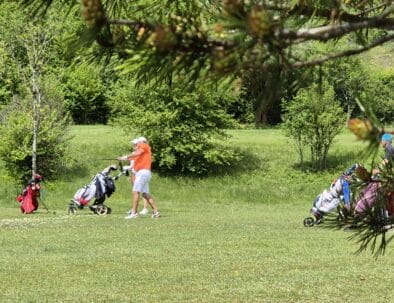 Un groupe de golfeurs sur un parcours verdoyant, profitant d'une journée ensoleillée. Un joueur, vêtu d'un t-shirt orange, s'apprête à frapper la balle avec son club, tandis que d'autres golfeurs se déplacent avec leurs chariots de golf. L'environnement est naturel, avec des arbres verdoyants en arrière-plan, créant une ambiance relaxante et propice à la pratique du golf. Cette image illustre parfaitement le plaisir de