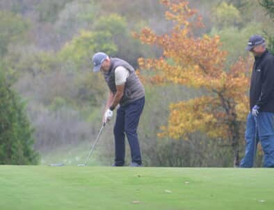 Un golfeur s'apprête à frapper la balle sur un terrain de golf en automne, entouré de feuillages colorés. Il porte un gilet et une casquette, montrant son engagement envers le sport. À l'arrière-plan, on aperçoit un autre joueur observant la scène. L'image capture l'essence d'une journée de golf dans un cadre naturel apaisant.