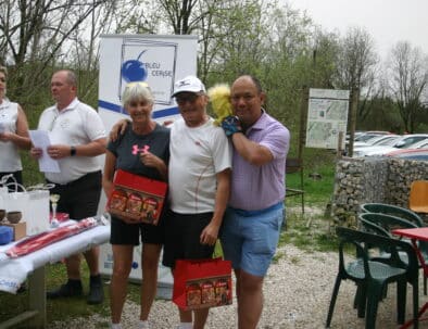 Cérémonie de remise de prix lors d'un événement organisé par Bleu Cerise. Sur l'image, des participants souriants reçoivent des récompenses, entourés de décorations et d'équipements sportifs. En arrière-plan, on aperçoit un panneau d'information et des voitures stationnées. L'événement met en avant la convivialité et le partage, avec des membres de l'organisation interagissant avec les participants.