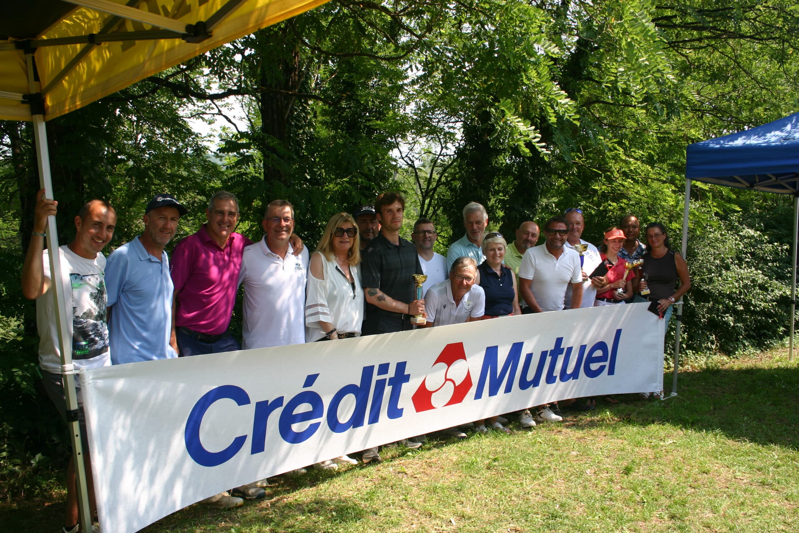 Groupe de personnes posant devant une banderole du Crédit Mutuel lors d'un événement en plein air, entouré d'arbres et d'une ambiance conviviale. L'image illustre l'engagement communautaire de la banque dans des activités locales.