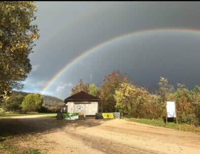Paysage naturel avec un arc-en-ciel vibrant au-dessus d'un bâtiment en pleine nature. Le ciel est partiellement nuageux, créant un contraste saisissant avec les couleurs de l'arc-en-ciel. Autour, des arbres aux feuilles dorées et vertes signalent l'automne, tandis qu'un chemin en terre mène à l'entrée du site. Cette image évoque la beauté des paysages extérieurs et les phénomènes météorologiques. Idéal pour les amateurs de