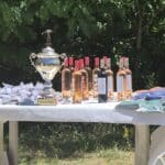 Table décorée pour un événement avec un trophée brillant au centre. À proximité, plusieurs bouteilles de vin rosé et rouge sont exposées, accompagnées de petits sachets et de divers articles en tissu, probablement des récompenses pour les participants. L'arrière-plan verdoyant ajoute une touche naturelle à l'ensemble, suggérant un cadre festif en plein air.