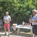 Un groupe de personnes assiste à une cérémonie en plein air, probablement pour la remise de prix après un tournoi. Deux hommes en tenues polo se tiennent devant une table décorée de coupes et de bouteilles de vin, suggérant un événement convivial lié au sport ou à une compétition. L'atmosphère est festive, entourée de verdure, idéale pour les rassemblements communautaires. Des participants se détendent sur des chaises, impliqués dans des conversations.