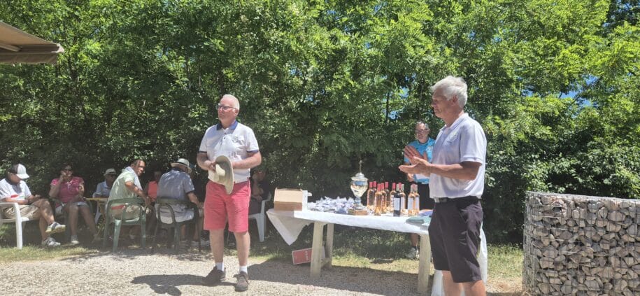 Un groupe de personnes assiste à une cérémonie en plein air, probablement pour la remise de prix après un tournoi. Deux hommes en tenues polo se tiennent devant une table décorée de coupes et de bouteilles de vin, suggérant un événement convivial lié au sport ou à une compétition. L'atmosphère est festive, entourée de verdure, idéale pour les rassemblements communautaires. Des participants se détendent sur des chaises, impliqués dans des conversations.