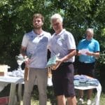 Cérémonie de remise de prix lors d'un tournoi de golf, deux participants posent ensemble en tenant leurs trophées. En arrière-plan, une table avec des récompenses et des sachets, entourée de verdure. Un organisateur lit les résultats, soulignant l'importance de l'événement sportif. Moment convivial célébrant l'esprit de compétition et la camaraderie entre joueurs.