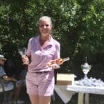 Une femme souriante en tenue de sport rose tient un verre et une bouteille de vin rosé lors d'un événement. En arrière-plan, des spectateurs assis, appréciant l'activité, tandis qu'un homme en polo bleu remet des prix. La scène se déroule en plein air, entourée de verdure, mettant en valeur l'atmosphère festive d'un tournoi ou d'une réunion sociale. Cette image illustre la convivialité et le plaisir partagés lors d'événements communautaires