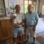 Deux hommes posent dans un clubhouse de golf après une compétition. L'un, tenant un trophée brillant portant le nom de Roland Perrin, exécute un sourire satisfait. L'autre homme, à ses côtés, porte une chemise bleu clair et des lunettes. En arrière-plan, des affiches de golf et un bar en bois créent une ambiance conviviale. Cet événement souligne la passion pour le golf et les échanges amicaux entre les joueurs.