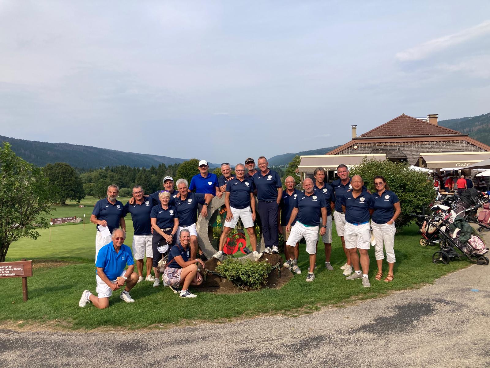 Groupe de golfeurs posant sur un terrain de golf verdoyant, vêtus de polos bleus et de shorts blancs. L'image montre une équipe souriante, rassemblée autour d'une sculpture en pierre, avec un paysage de collines en arrière-plan. Des chariots de golf sont visibles à droite, et un club-house se trouve en arrière-plan. Cette photo évoque une journée conviviale dédiée au golf, idéale pour mettre en avant des événements sportifs ou des rencontres am