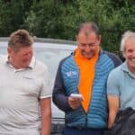 Quatre hommes souriants se tiennent côte à côte sur un terrain de golf, chacun tenant un sac de golf. L'un des hommes, vêtu d'une veste bleue et d'un t-shirt orange, consulte son téléphone. En arrière-plan, un véhicule est stationné près d'un drapeau portant le logo d'une marque. Cette image évoque un moment convivial, probablement après un tournoi de golf, mettant en avant l'esprit d'équipe et l'amitié dans le cadre d'activ