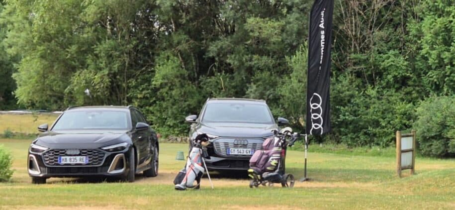 Deux voitures Audi élégantes sont garées sur un terrain de golf verdoyant, avec un drapeau Audi flottant à proximité. À côté des voitures, un chariot de golf est visible, prêt pour une partie. L'environnement naturel, riche en arbres, crée une ambiance sereine et propice au golf. Cette image illustre le luxe et le style de vie associé à la pratique de ce sport.