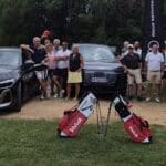 Groupe de golfeurs posant fièrement devant deux voitures Audi lors d'un événement sportif. Les participants, vêtus de tenues de golf colorées, affichent un esprit convivial et détendu. En arrière-plan, un drapeau avec l'inscription "Nous sommes..." et des sacs de golf Volvik ajoutent une touche professionnelle à l'image. Cette scène illustre l'esprit de camaraderie et le plaisir partagé autour du golf.