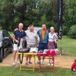 Groupe de golfeurs posant sur un terrain de golf, avec une voiture Audi en arrière-plan. Ils se tiennent près d'une table chargée d'équipements de golf, dont des sacs Volvik et divers accessoires. L'environnement est verdoyant, typique d'un parcours de golf, et l'ambiance est conviviale. Cette image illustre un événement de golf, mettant en avant la passion pour ce sport et les rencontres entre amateurs.