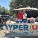 Image d'une remise de prix lors d'un événement sportif, avec des participants posant devant une table décorée du logo Hyper U Champagnole. Sur la table, on peut voir des récompenses, y compris un appareil Moulinex. En arrière-plan, des spectateurs applaudissent et des golfeurs se préparent à jouer. L'événement se déroule en plein air, sous un ciel ensoleillé, créant une ambiance conviviale et festive.