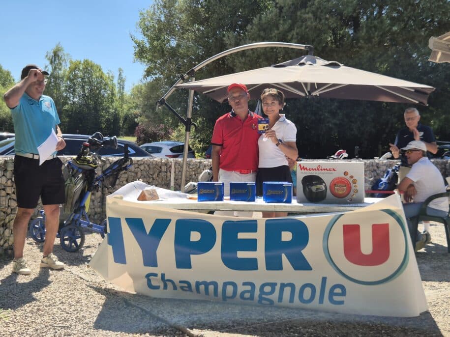 Image d'une remise de prix lors d'un événement sportif, avec des participants posant devant une table décorée du logo Hyper U Champagnole. Sur la table, on peut voir des récompenses, y compris un appareil Moulinex. En arrière-plan, des spectateurs applaudissent et des golfeurs se préparent à jouer. L'événement se déroule en plein air, sous un ciel ensoleillé, créant une ambiance conviviale et festive.