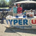 Deux hommes souriants posent devant un stand décoré aux couleurs de Hyper U Champagnole lors d'un événement en plein air. Le stand présente des produits, dont un robot culinaire Moulinex, et des prix. En arrière-plan, des golfeurs et des équipements de golf sont visibles, évoquant une journée conviviale dédiée au golf. L'ambiance est festive, avec un ciel ensoleillé et des espaces verts. Cette image illustre l'engagement de Hyper U dans