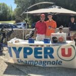 Deux participants souriants posent devant un stand marqué "HYPER U Champagnole" lors d'un événement en plein air. Le stand présente des produits comme un autocuiseur Moulinex et des barres énergétiques. À l'arrière-plan, des golfeurs se préparent à jouer, tandis que d'autres spectateurs se détendent sur des chaises. L'image capture l'atmosphère conviviale d'une journée de golf estivale.