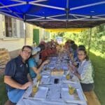 Groupe de personnes réunies sous un grand barnum pour un repas convivial en extérieur. La table est dressée avec des assiettes blanches et des verres, tandis que des mets appétissants sont visibles. L'ambiance est détendue, propice aux échanges et à la convivialité, entourée par la verdure. Cette image illustre un moment de partage et de célébration, parfait pour des événements en plein air.