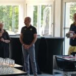 Événement convivial avec une intervenante prenant la parole, accompagnée de deux hommes, l'un en tenue sportive et l'autre tenant des livres. En arrière-plan, des verres à vin sont soigneusement disposés sur une table, tandis que de grandes fenêtres laissent entrer la lumière naturelle. Cette scène se déroule dans un cadre moderne, propice aux rassemblements et aux échanges.