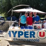 Un événement de golf se déroule à Champagnole, avec des participants rassemblés autour d'une table sponsorisée par Hyper U. Trois hommes, souriants, posent ensemble avec des prix, tandis que d'autres participants profitent de l'atmosphère conviviale. En arrière-plan, on aperçoit des sacs de golf et des voitures. La scène évoque une journée ensoleillée de compétition amicale, illustrant le plaisir du golf et de la camaraderie.