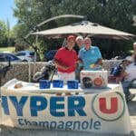 Un événement convivial de golf se déroule en plein air, avec des participants rassemblés autour d'une table décorée aux couleurs de Hyper U Champagnole. Deux hommes, souriants, posent avec des prix, dont un robot de cuisine Moulinex, tandis qu'un autre participant consulte des documents. En arrière-plan, des golfeurs se préparent à jouer, créant une ambiance dynamique et festive. L'événement met en avant des activités sportives et des interactions communautaires.