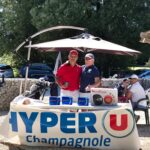 Événement de golf à Champagnole avec des participants réunis autour d'une table sponsorisée par Hyper U. Les golfeurs, vêtus de polos colorés, interagissent dans une ambiance conviviale. Des équipements de golf sont visibles en arrière-plan, ainsi que des prix sur la table. Le cadre extérieur est agréable, avec des arbres et des parasols, créant une atmosphère détendue pour les amateurs de golf.