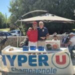 Deux hommes souriants posent ensemble devant une table décorée avec un logo HYPER U à Champagnole. Ils tiennent des récompenses, tandis qu'une variété de prix, dont un appareil Moulinex, est exposée sur la table. En arrière-plan, des golfeurs et des équipements de golf sont visibles, soulignant l'ambiance conviviale d'un événement sportif en plein air. Le ciel est dégagé, suggérant une journée ensoleillée idéale pour jouer au