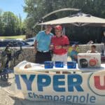 Image d'un événement de golf à Champagnole, mettant en avant un stand de promotion avec des produits de la marque Moulinex. Deux hommes, l'un en polo bleu et l'autre en polo rouge, interagissent autour d'une table où sont exposés des articles. En arrière-plan, des participants assistent à l'événement, et des voitures sont garées à proximité. L'atmosphère est ensoleillée et conviviale, idéale pour un rassemblement sportif.
