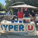 Un événement convivial se déroule à Champagnole, avec des participants se rassemblant autour d'un stand de l'enseigne Hyper U. Deux hommes posent devant un comptoir, mettant en avant des produits, dont un robot de cuisine Moulinex. En arrière-plan, plusieurs golfeurs se préparent, soulignant l'atmosphère sportive et festive de la journée. Le cadre ensoleillé et verdoyant contribue à l'ambiance agréable de cette rencontre autour du golf.