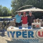 Un groupe de golfeurs posant ensemble lors d'un événement en plein air, avec un stand de la marque Hyper U Champagnole visible en arrière-plan. Des prix, comme un multicuiseur Moulinex et d'autres articles, sont exposés sur une table. Environnement ensoleillé avec des voitures stationnées et des arbres verdoyants autour, créant une ambiance conviviale et festive.