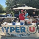 Événement de golf organisé par Hyper U Champagnole, avec des participants souriants autour d'une table décorée. Le stand présente des produits, dont un autocuiseur Moulinex, et des échantillons de la marque Sunquest. En arrière-plan, on aperçoit des golfeurs et des équipements de golf. Une journée ensoleillée propice aux échanges et à la convivialité dans un cadre naturel.