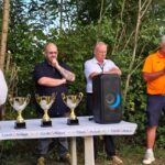 Cérémonie de remise de trophées en plein air avec plusieurs intervenants. Quatre coupes sont exposées sur une table décorée aux couleurs du Crédit Mutuel. Les participants, habillés de manière décontractée, échangent des discours devant un public. L'événement se déroule dans un cadre naturel, entouré d'arbres. Ambiance conviviale et festive, idéale pour célébrer les réussites sportives.
