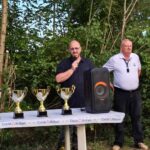 Cérémonie de remise des trophées en plein air, avec plusieurs intervenants sur scène. Un homme prend la parole avec un microphone, tandis que d'autres membres de l'équipe se tiennent à ses côtés. Sur la table, plusieurs coupes et trophées brillants sont exposés, symbolisant les réussites des participants. En arrière-plan, une végétation luxuriante complète le cadre de cet événement festif.