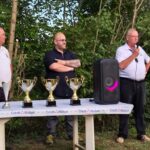 Cérémonie de remise de prix en plein air avec plusieurs intervenants. Quatre trophées sont exposés sur une table, tandis que les orateurs, vêtus de polos et de chemises, s'adressent à un public en arrière-plan. L'événement se déroule dans un cadre naturel, entouré de verdure, et un haut-parleur est présent sur la table pour l'animation. Cette image illustre un moment festif et communautaire, mettant en avant la