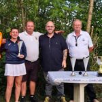 Cérémonie de remise de prix de golf avec des participants souriants, dont une femme tenant un trophée, entourée de plusieurs hommes. Ils se tiennent devant une table avec des coupes dorées et un fond verdoyant. L'événement est organisé par le Crédit Mutuel, mettant en avant l'esprit sportif et la camaraderie.