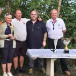 Cérémonie de remise de prix avec des participants souriants, tenant des trophées sur une table décorée. L'événement se déroule en plein air, entouré de verdure, et met en avant l'esprit de compétition et de camaraderie. Les trophées symbolisent les réalisations des gagnants dans une compétition locale. L'atmosphère est festive, reflétant la passion pour le sport et les activités communautaires.