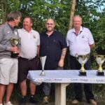 Cérémonie de remise de trophées après un événement sportif, avec des participants souriants et des coupes sur une table. Les gagnants sont entourés d'organisateurs, soulignant l'esprit de camaraderie et de compétition. L'arrière-plan verdoyant ajoute une touche naturelle à l'événement, tandis qu'un haut-parleur indique une ambiance festive. Cette image illustre l'importance des compétitions locales et de la reconnaissance des performances sportives.