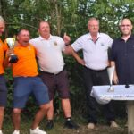 Cérémonie de remise de trophées avec des participants souriants, tous vêtus de t-shirts orange. Un homme au centre, tenant un trophée, célèbre sa victoire entouré de ses coéquipiers et officiels. Sur la table, plusieurs coupes témoignent des récompenses attribuées lors de l'événement. En arrière-plan, une verdure luxuriante crée une atmosphère festive pour cette compétition sportive.