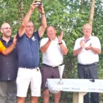 Cinq hommes se tiennent ensemble lors d'une cérémonie de remise de prix en extérieur, entourés de verdure. L'un d'eux, vêtu d'un polo bleu, lève un trophée en signe de victoire, tandis que les autres applaudissent et expriment leur joie. L'événement semble célébrer un succès sportif, avec un décor naturel qui ajoute une ambiance conviviale. Une table avec un haut-parleur et des éléments de branding de Crédit Mutuel est visible à l