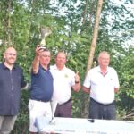 Cérémonie de remise de trophée en plein air, avec quatre hommes souriants posant ensemble. L'un des participants, portant une chemise bleue, soulève fièrement un trophée. Les autres membres du groupe affichent des expressions de joie et d'encouragement. À l'arrière-plan, une verdure luxuriante crée une atmosphère naturelle. Sur la table devant eux, on peut apercevoir un haut-parleur et un décor évoquant un événement