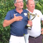 Cérémonie de remise de trophée lors d'un événement sportif, avec un homme exprimant sa joie tout en tenant un trophée. En arrière-plan, des arbres verdoyants créent une ambiance naturelle. Un autre participant, souriant, se tient à ses côtés. La table est ornée d'une nappe avec le logo d'un sponsor, Crédit Mutuel, soulignant le soutien d'entreprises locales aux activités sportives. Cette image illustre l'esprit de camaraderie et