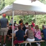 Groupe de jeunes participants assis sous un chapiteau, attentifs à une présentation en plein air. L'événement se déroule dans un cadre naturel verdoyant, avec des adultes et des enfants portant des casquettes colorées. L'ambiance est conviviale et propice à l'apprentissage, suggérant une activité éducative ou sportive. Les tables en bois et les chaises blanches ajoutent à l'atmosphère décontractée de la rencontre.