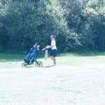 Un golfeur marche sur un parcours verdoyant, poussant une voiture de golf. L'environnement est paisible, entouré d'arbres verdoyants. La scène évoque une journée ensoleillée, idéale pour jouer au golf. L'individu porte une tenue sportive adaptée, soulignant l'aspect dynamique de l'activité. Cette image reflète l'importance du golf comme loisir en plein air.