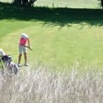 Une jeune golfeuse s'apprête à frapper une balle sur le green d'un parcours de golf ensoleillé. Elle porte un polo rose et un short, avec un chapeau pour se protéger du soleil. À proximité, un chariot de golf est garé, contenant divers clubs. L'arrière-plan est composé de verdure, mettant en valeur l'environnement paisible et naturel du golf. Cette image illustre la passion pour le golf et la pratique du sport en plein air.