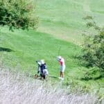 Golfeur en action sur un parcours verdoyant, préparant son swing avec un club de golf. Un caddy avec un chariot est visible à proximité. L'environnement naturel est agrémenté d'arbres et d'herbes sèches, illustrant une journée ensoleillée propice au golf. Cette image évoque la passion pour le sport et la beauté des terrains de golf.