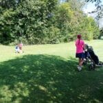 Deux enfants jouent sur un terrain de golf verdoyant. L'un est accroupi pour ramasser un objet, tandis que l'autre, portant un chariot de golf, marche sur l'herbe bien entretenue. L'environnement est ensoleillé, avec des arbres en arrière-plan, offrant une ambiance agréable pour une journée d'activité en plein air. Cette scène illustre l'importance du golf comme activité familiale et de loisirs, favorisant le plaisir et le sport en nature.