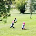 Trois golfeuses en tenue colorée, dont deux en rose, jouent sur un parcours verdoyant. Elles utilisent des chariots de golf pour transporter leurs clubs. L'environnement est paisible, avec des arbres et une lumière naturelle agréable, typique d'une journée ensoleillée sur le golf. Cette scène illustre l'activité de golf en plein air, favorisant le sport et la détente en nature.